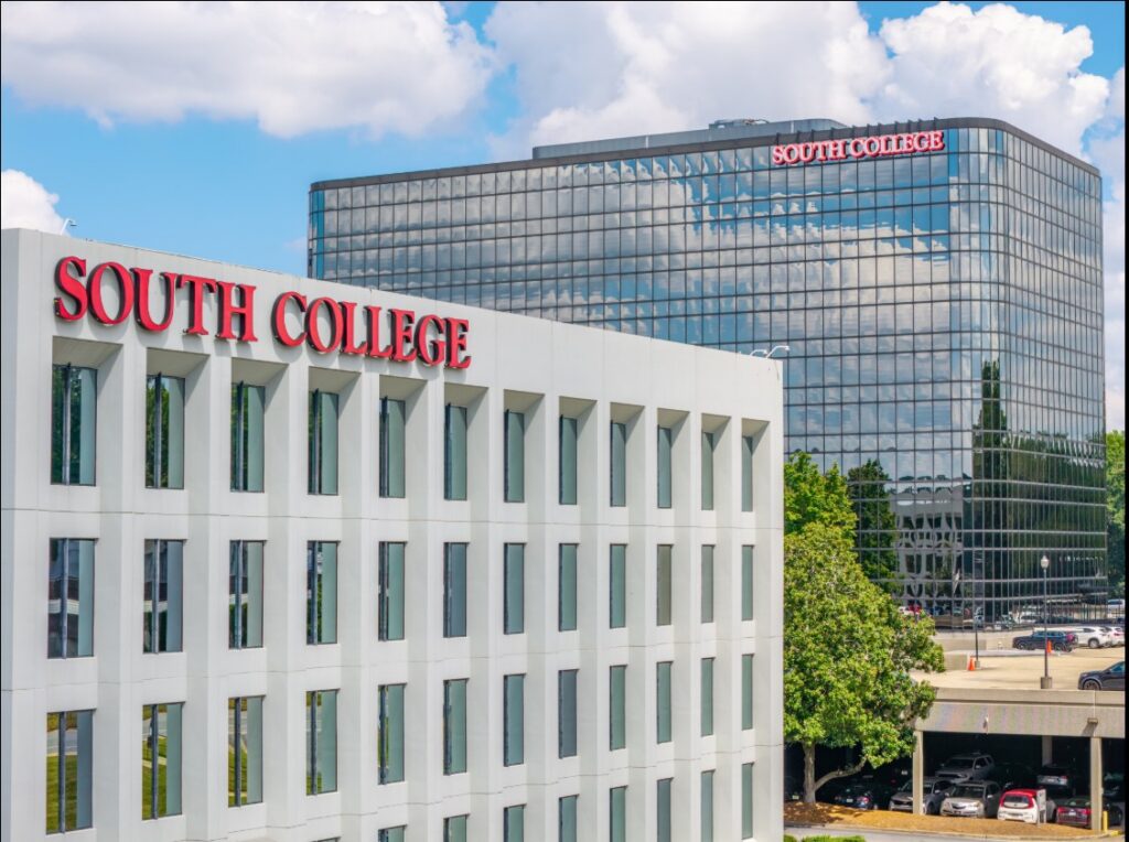 Two modern buildings with “South College” in red letters on each. The foreground building is white and rectangular, while the taller glass building in the background reflects the sky and clouds. Trees and parked cars are visible.