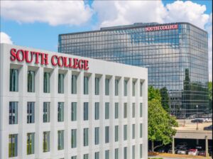 Two modern buildings with “South College” in red letters on each. The foreground building is white and rectangular, while the taller glass building in the background reflects the sky and clouds. Trees and parked cars are visible.