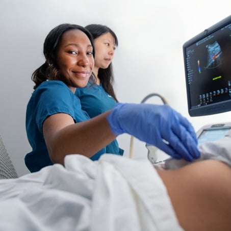 Two imaging science students conduct an ultrasound examination. One person, wearing blue scrubs and gloves, smiles at the camera while holding the ultrasound probe on a patient's abdomen. The other person, also in blue scrubs, monitors the ultrasound screen.