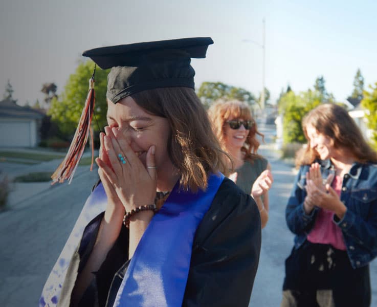 A graduate wearing a cap and gown covers her face with her hands, appearing emotional. Two students in the background are clapping and smiling, celebrating her achievement on a sunny day.