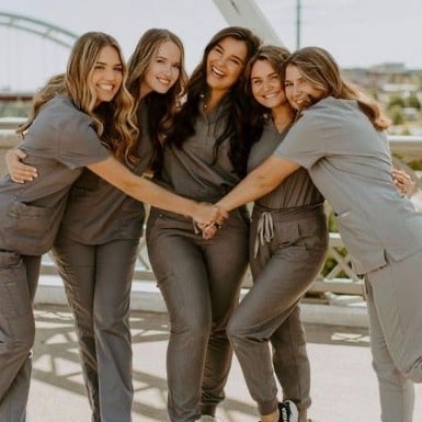 Five female students in matching gray scrubs stand close together on a sunny outdoor bridge. They are smiling and hugging each other warmly, radiating happiness and camaraderie. A scenic urban background with a bridge structure is visible in the distance.
