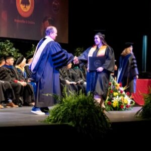A graduate in a cap and gown shakes hands with the Chancellor on stage during a graduation ceremony. Other faculty members are seated in the background, and there's a floral arrangement nearby.