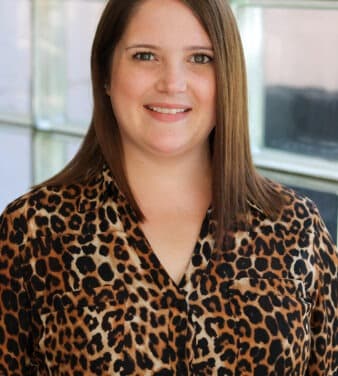 A woman with straight brown hair wearing a leopard print blouse stands in front of a window, smiling at the camera.