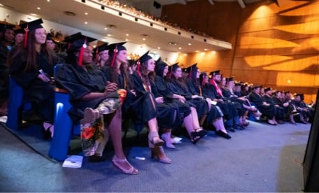A group of graduates in caps and gowns sit in rows on stage during a commencement ceremony in an auditorium. The audience is visible in the background.