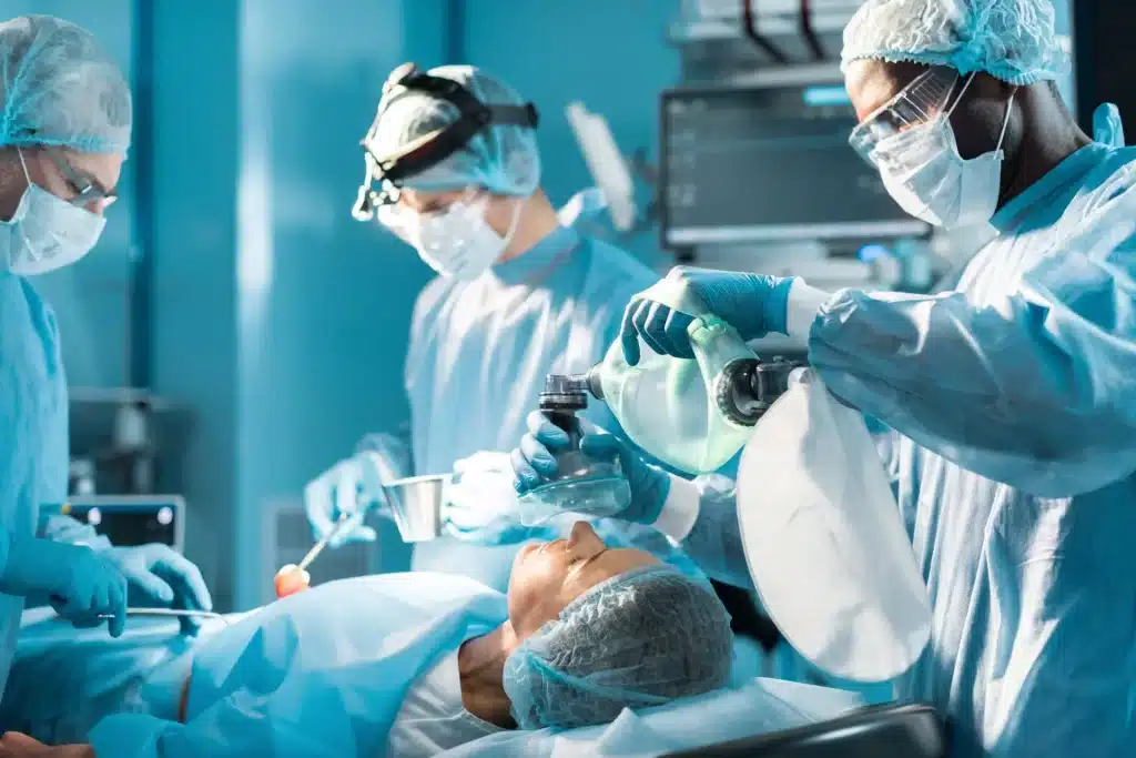 A student surgical technologist team operates on a patient in an operating room. The patient is lying on the operating table with an oxygen mask placed on their face by one of the students. The team is focused, wearing scrubs, masks, gloves, and surgical caps.