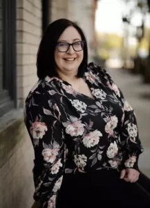 A woman with shoulder-length dark hair and glasses is smiling while leaning against a brick wall outdoors. She is wearing a black floral blouse.