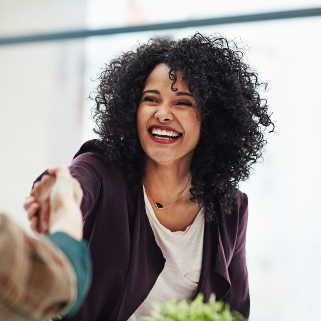 A person with curly hair is smiling brightly while shaking hands with someone out of frame. They are wearing a dark blazer over a light top, and there is a small houseplant in the foreground. The background is out of focus, giving a warm, cheerful vibe.