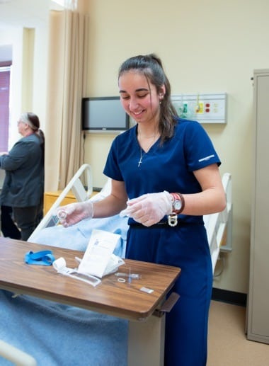 A nursing student in blue scrubs stands at a patient's bedside table, smiling and preparing medical supplies. She wears gloves and various wristbands.