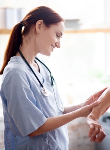 A nursing student with a stethoscope around her neck is smiling as she examines a patient's arm. She is wearing a light blue medical scrub top. The background is softly lit, suggesting a bright medical environment.