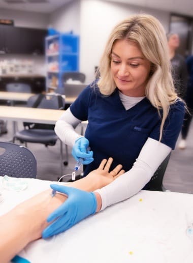 A woman wearing navy blue scrubs and blue gloves is practicing a medical procedure on a mannequin's arm. She appears focused, holding a needle and an IV line in a simulation lab setting with chairs and medical supplies in the background.