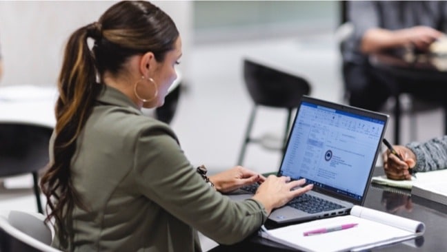 A woman with long hair tied in a ponytail works on a laptop at a desk in an office. The screen displays an email program.