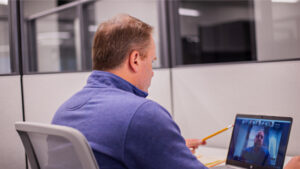 A man in a blue sweater, sitting in a modern office cubicle, participates in a virtual meeting on his laptop.