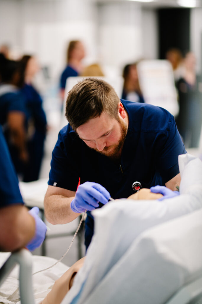 A medical professional wearing navy scrubs and gloves is focused on performing a procedure on a medical dummy. In the background, several blurred figures in similar attire are engaged in various activities, indicating a busy healthcare training environment.