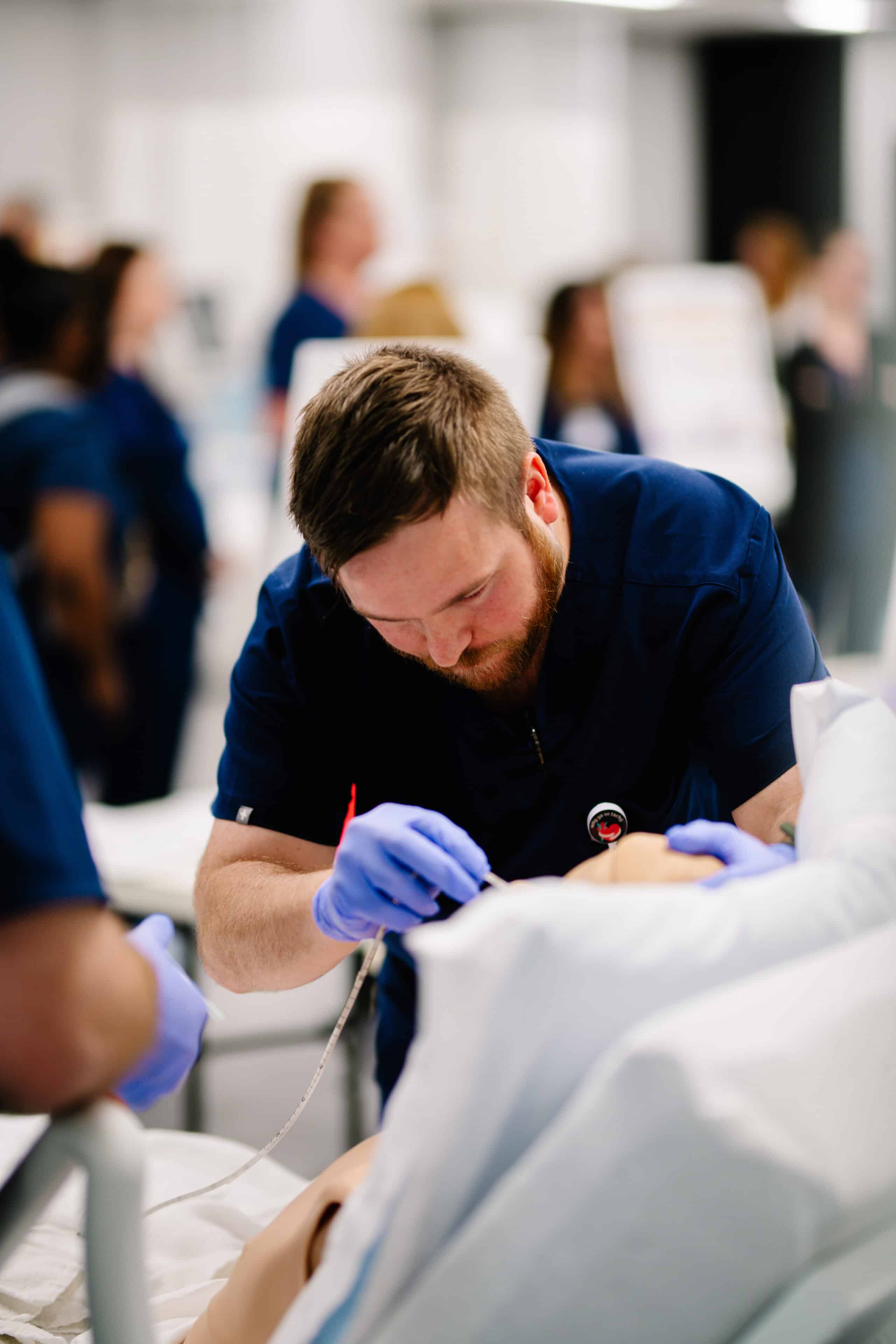 A medical professional wearing navy scrubs and gloves is focused on performing a procedure on a medical dummy. In the background, several blurred figures in similar attire are engaged in various activities, indicating a busy healthcare training environment.