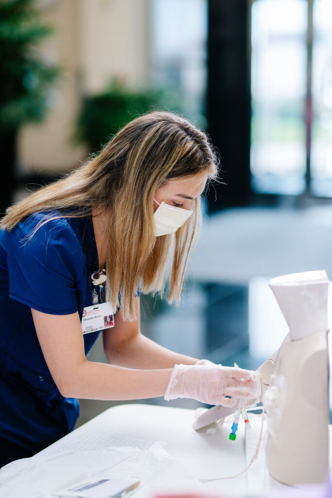 A healthcare professional wearing blue scrubs and a face mask is practicing on a medical dummy. They are closely focused, wearing gloves, and handle medical equipment, indicating a training or educational scenario. A clipboard with papers is visible on the table.