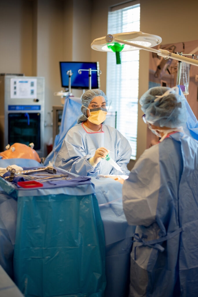 Two surgeons in blue gowns and face masks perform surgery in a well-lit operating room. Surgical instruments are laid out on a table covered with blue sterile drapes. One surgeon is focusing intently on the procedure while the other assists.