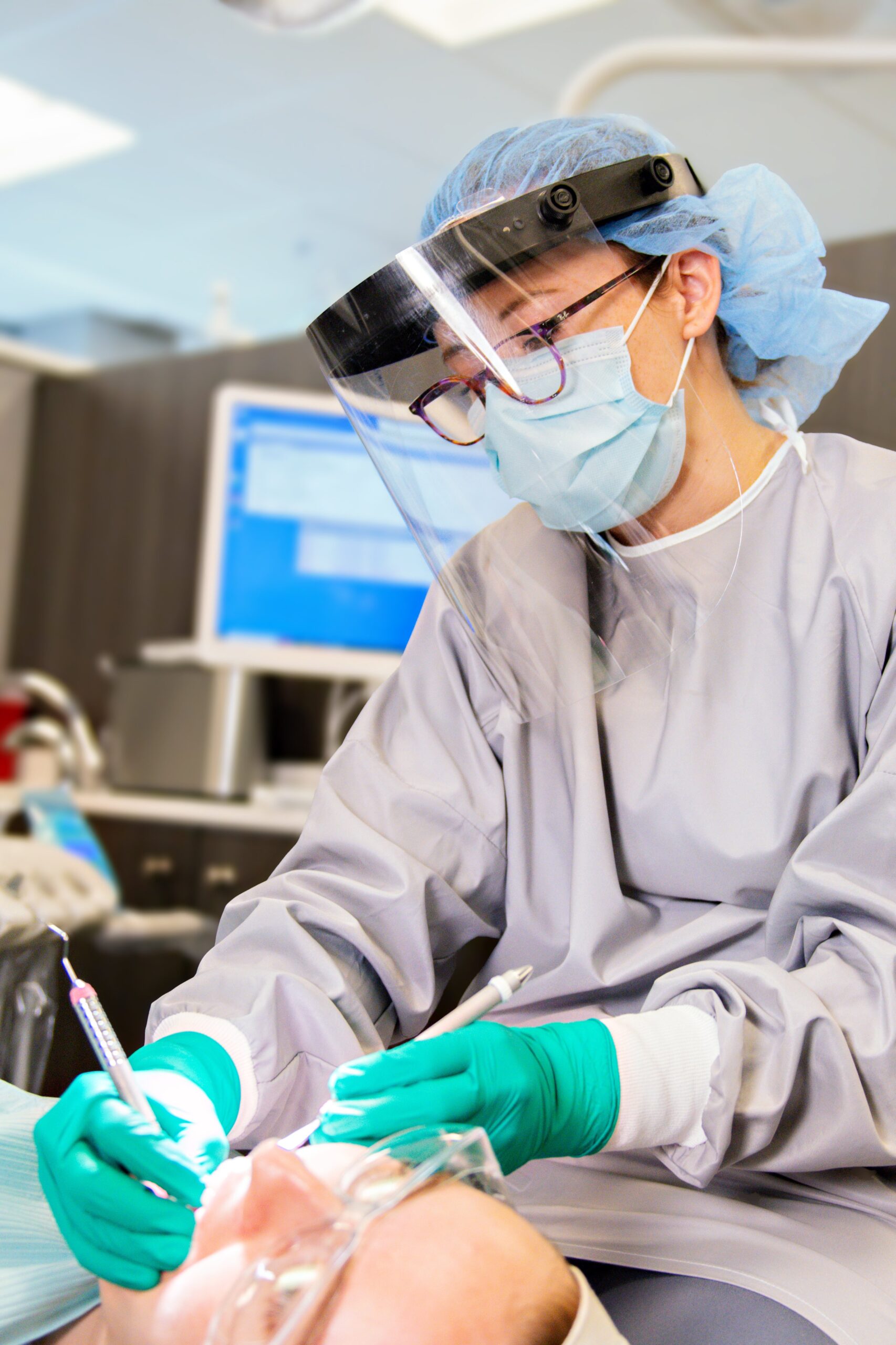 A dental student wearing protective gear, including a face shield, mask, gloves, and a gown, performs a dental procedure on a patient. The student holds a dental instrument while the patient lies back in the dental chair with their mouth open. A computer monitor is seen in the background.