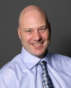 A man with a friendly smile is wearing a light blue dress shirt and a patterned tie. He is posed against a neutral gray background.