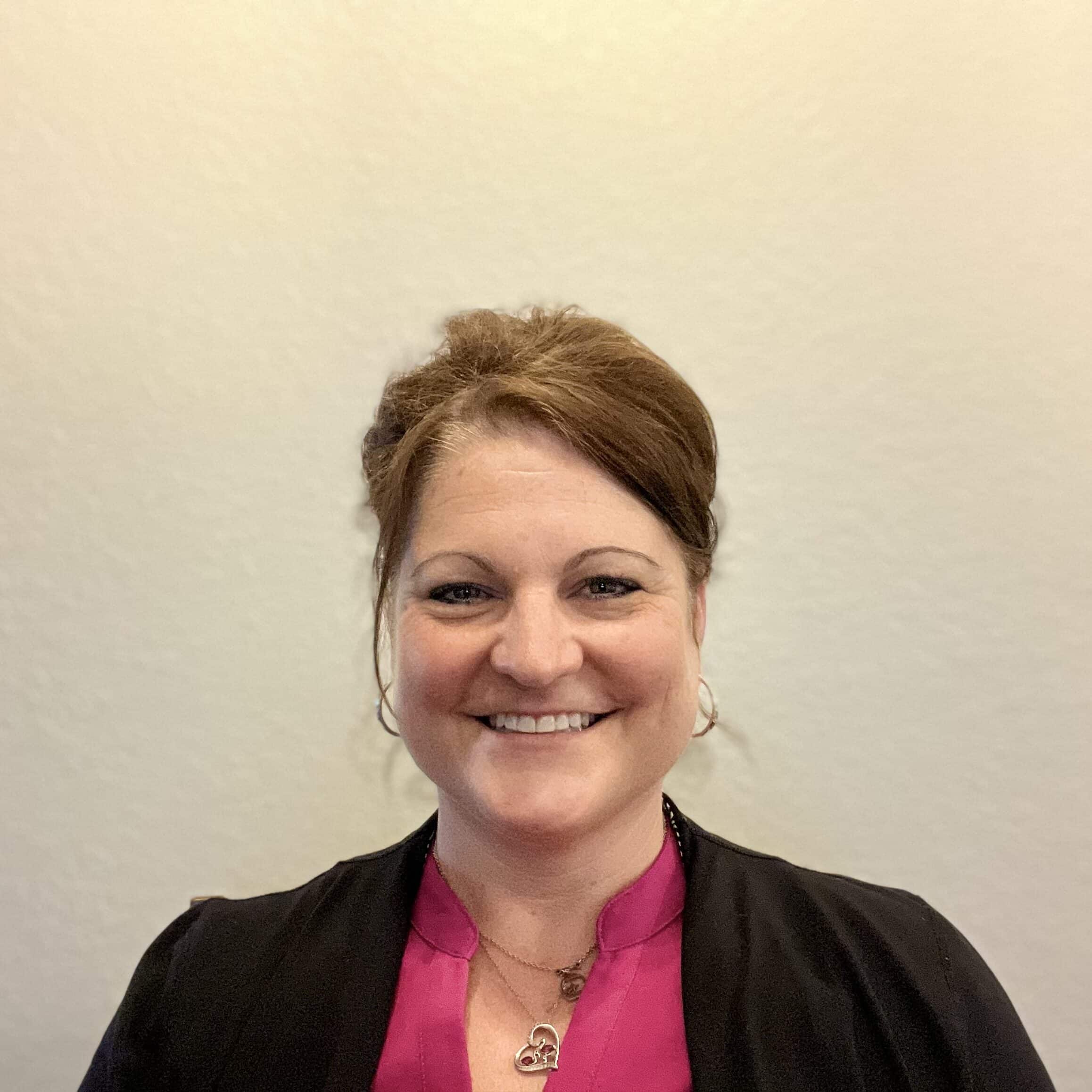 A person with brown hair styled up is smiling. They are wearing a pink top underneath a black jacket and hoop earrings. The background is a plain beige wall.