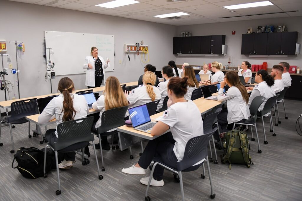A group of nursing students in white coats are seated at desks in a classroom, attentively listening to an instructor who is standing and speaking near a whiteboard. The students have laptops and notebooks in front of them. The room is equipped with medical supplies.