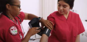 A healthcare professional in a red uniform checks a patient's blood pressure using a cuff in a medical setting. The patient is also wearing a red shirt.