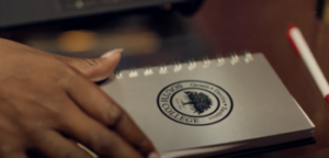 Close-up of a hand holding a small spiral-bound notebook with an emblem reading "Floo Hudson College." The notebook lies on a dark surface next to a red and white pen.