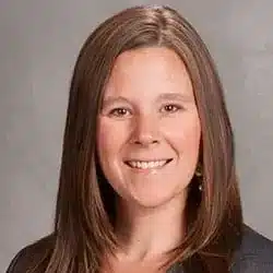 A woman with long, straight brown hair smiles at the camera. She is wearing a gray blazer over a pink top, and the background is a plain gray backdrop.