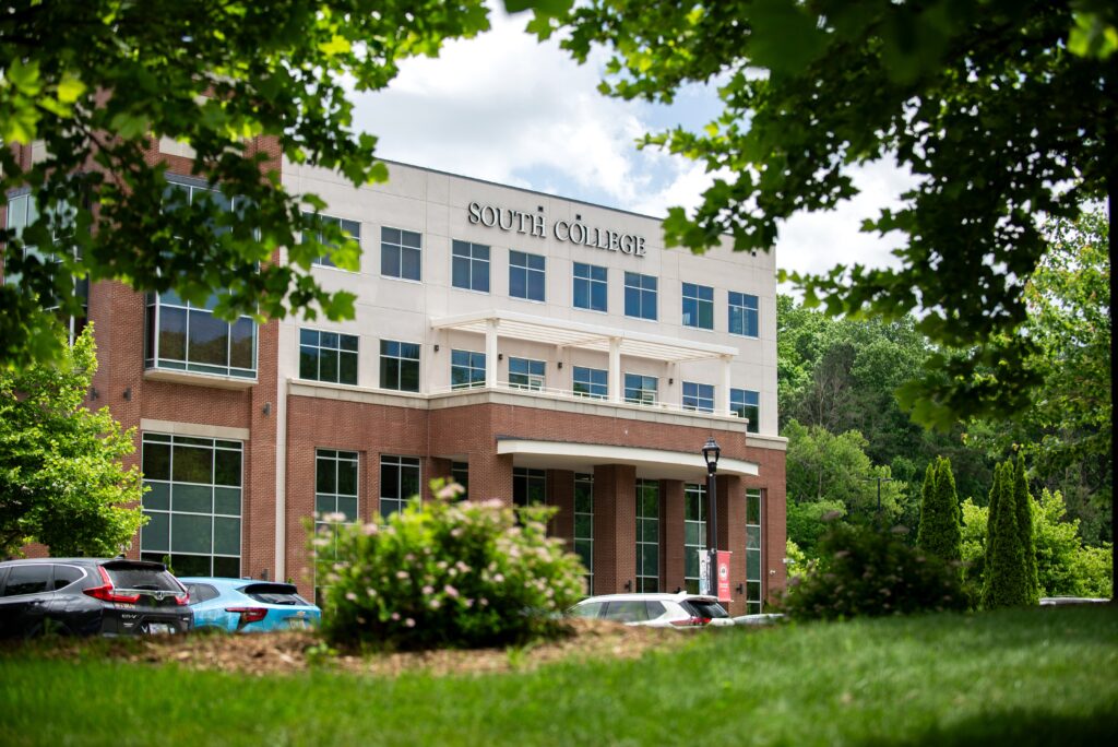 A modern brick and white building with "South College" signage on the front, surrounded by green trees and parked cars.