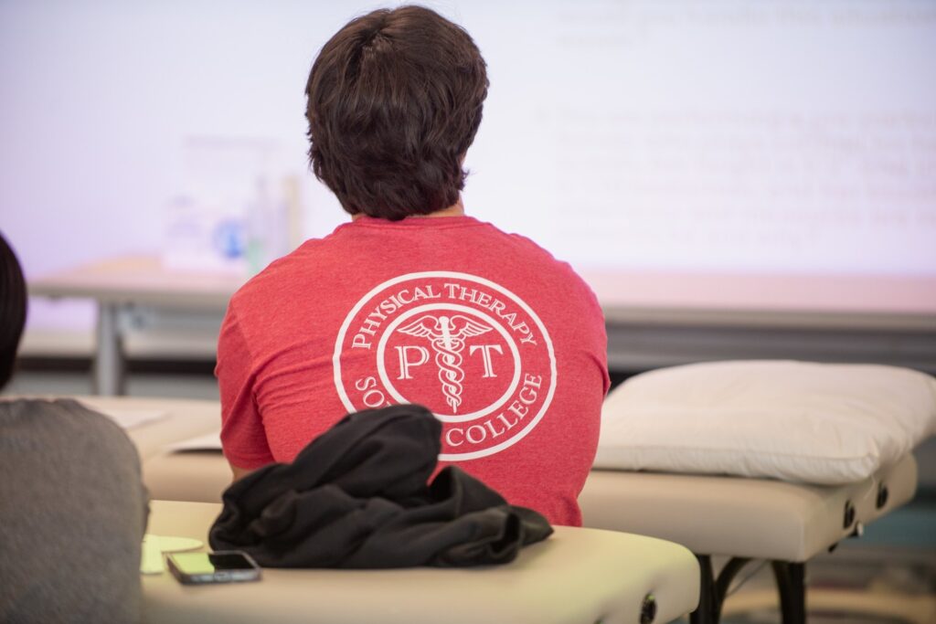 A person with dark hair wearing a red "Physical Therapy South College" shirt sits on a massage table, facing away from the camera in a classroom setting with a pillow and jacket nearby.