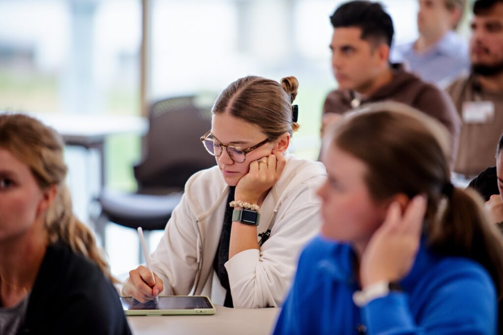 A group of students sits in a classroom. A woman in the center, wearing glasses and a white hoodie, writes on a tablet, appearing focused. Other students are seated around her, listening attentively.
