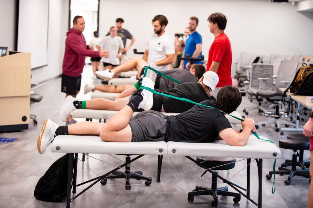 A group of people in athletic wear practice physical therapy techniques in a classroom, with some lying on treatment tables using resistance bands while others observe and participate.