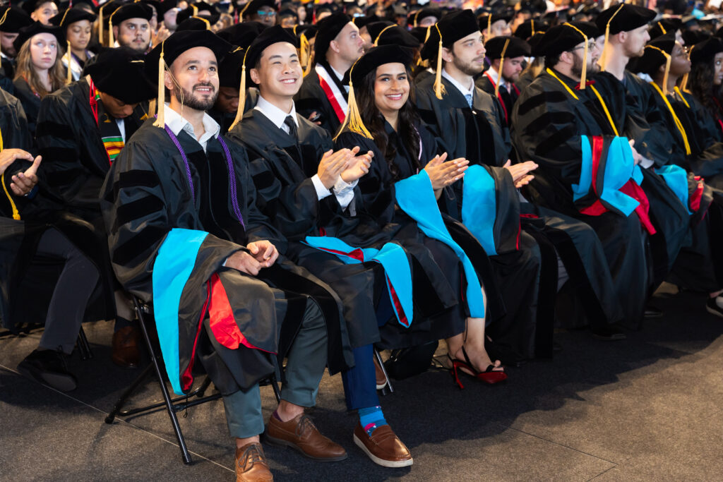 A group of graduates in black caps and gowns sit in rows at a ceremony, smiling and clapping. Colored sashes, tassels, and acknowledgments for financial aid are visible, adding to the celebratory atmosphere.