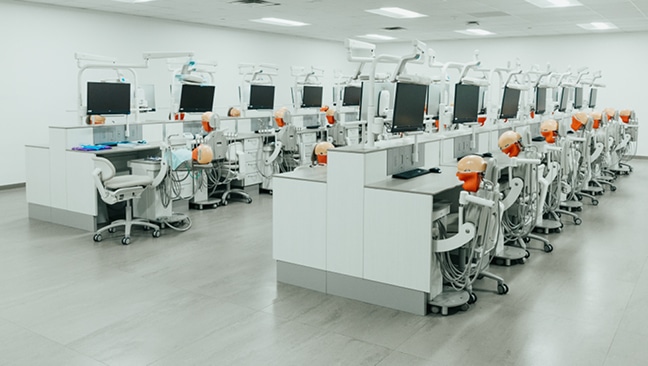 A modern dental training lab with rows of workstations, each equipped with computers, dental chairs, and realistic dental mannequins, all in a clean, bright room.