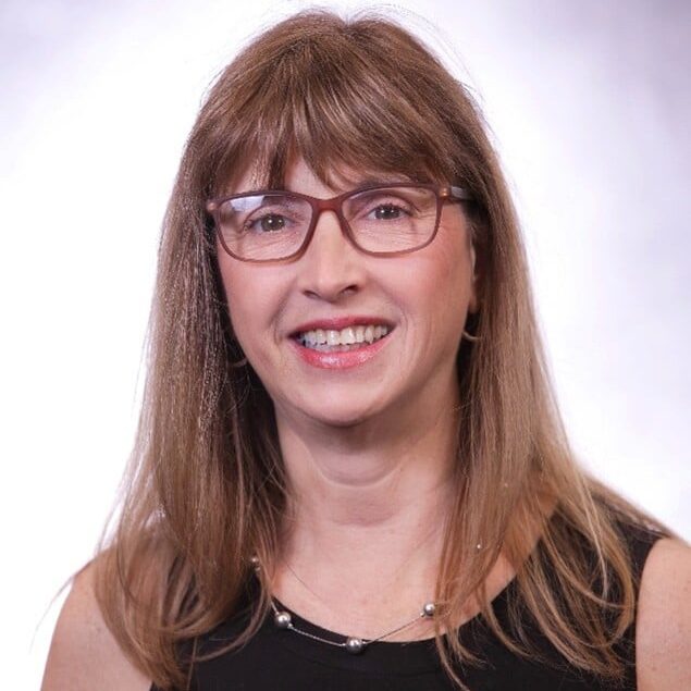 A woman with straight light brown hair, wearing glasses, a sleeveless black top, and a delicate necklace, smiles at the camera against a light, softly blurred background.