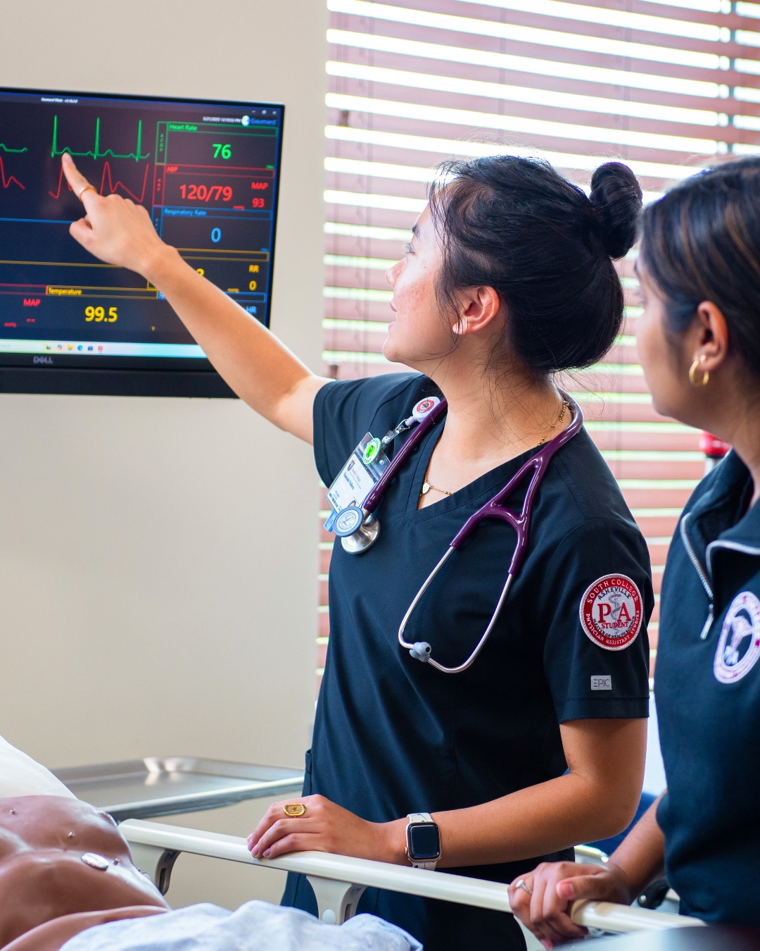 Two healthcare professionals in scrubs examine and discuss a monitor displaying a patient's vital signs and ECG in a clinical setting, with one pointing at the screen. A medical mannequin lies on the bed in front of them.