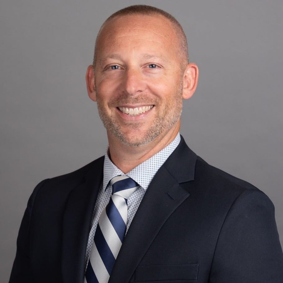 A smiling man with short hair and a trimmed beard wearing a dark suit, white patterned shirt, and a striped tie, posing against a plain gray background.