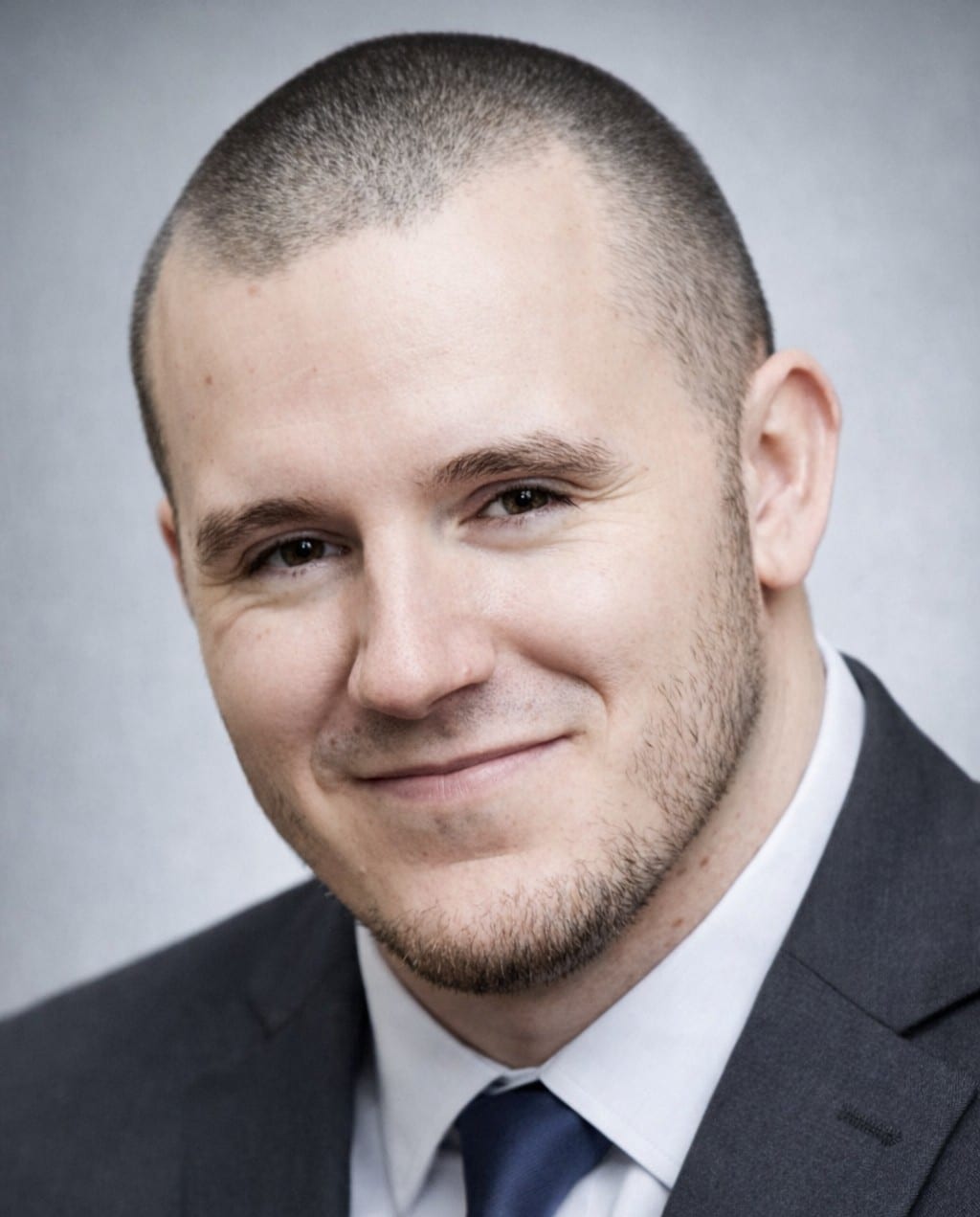 A man with a close-cropped haircut and light stubble is smiling slightly. He is wearing a dark suit jacket, white shirt, and dark tie, with a neutral gray background behind him.