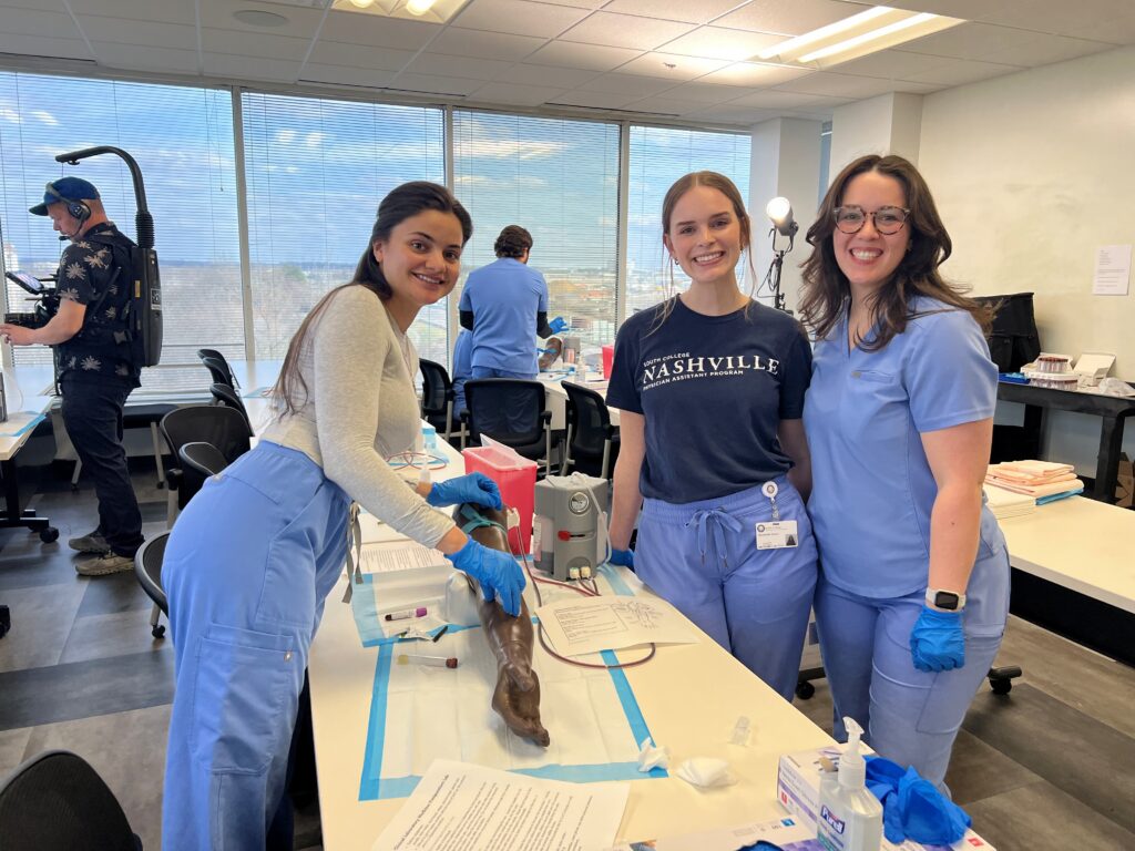 Three smiling women in scrubs stand around a medical training station with a mannequin arm, practicing clinical skills in a bright classroom. Two other people work in the background near large windows.
