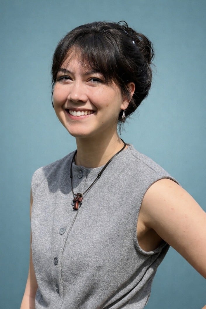 A young woman with dark hair in a bun smiles at the camera. She is wearing a sleeveless gray button-up top and a necklace with a pendant, standing against a plain blue background.