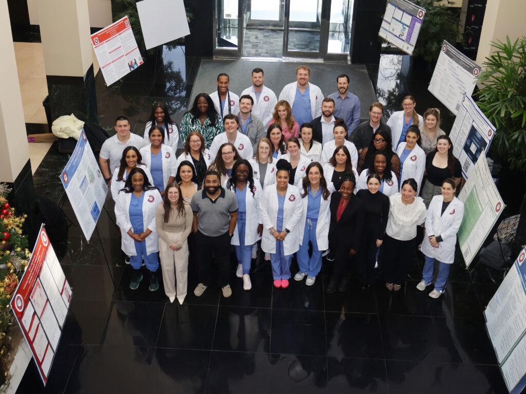 A large group of people, some in white lab coats and scrubs, stand together indoors, smiling at the camera. Behind them are scientific posters on display, suggesting a professional or academic event.