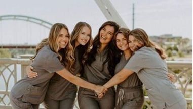 Five women in gray scrubs stand closely together on a bridge, smiling and hugging each other. The background shows an arched bridge and a cityscape under a clear sky.