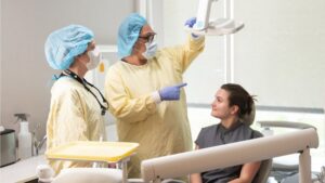 Two dental professionals in protective gowns and masks prepare a dental X-ray machine while a patient sits in the chair, looking up at them and smiling.