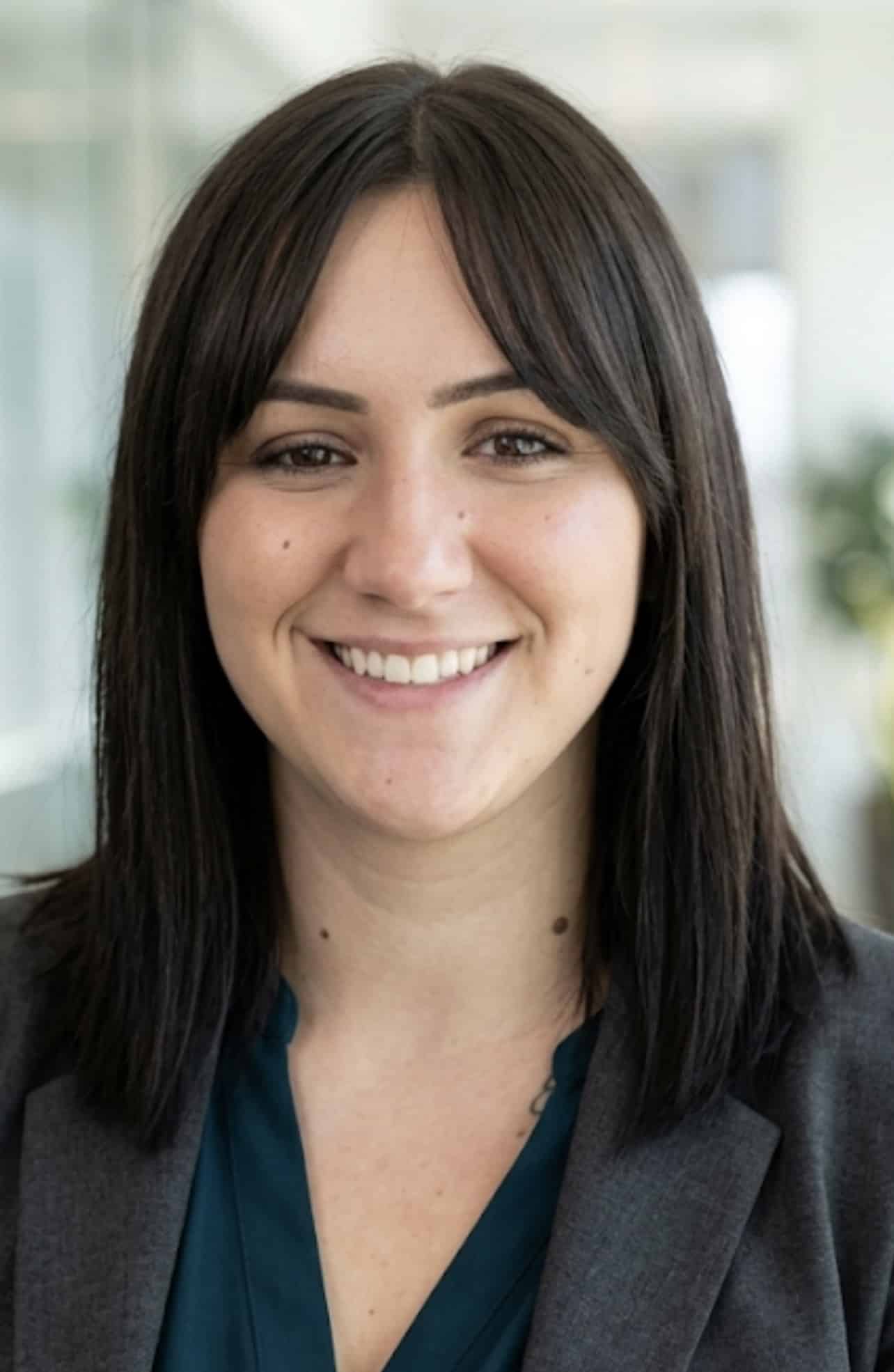 A woman with straight dark hair and bangs, wearing a teal blouse and gray blazer, smiles in a brightly lit office setting.