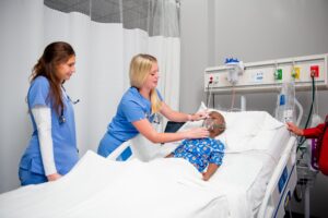 Two nurses in blue scrubs attend to a child in a hospital bed, adjusting an oxygen mask on the patient, surrounded by medical equipment and white curtains.