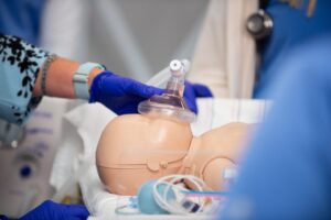 A person wearing blue gloves holds an oxygen mask over a medical mannequin’s face during a simulated medical procedure, with medical equipment visible nearby.